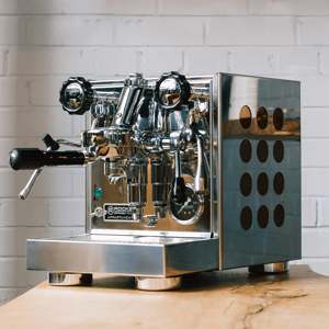 Rocket Appartamento stainless-steel espresso machine with E61 group head, portafilter, pressure gauge, and circular side cut-outs on a wooden counter.